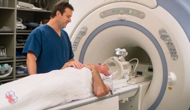 A specialist stands by an MRI machine as a person lays on a platform outside the MRI machine.